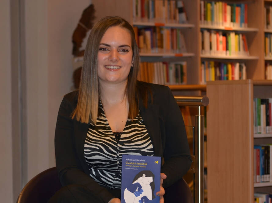 Ritratto fotografico di Valentina Giacalone seduta in una libreria, che sorride tenendo in mano una copia del suo libro dalla copertina blu, intitolato "Cicatrici invisibili".